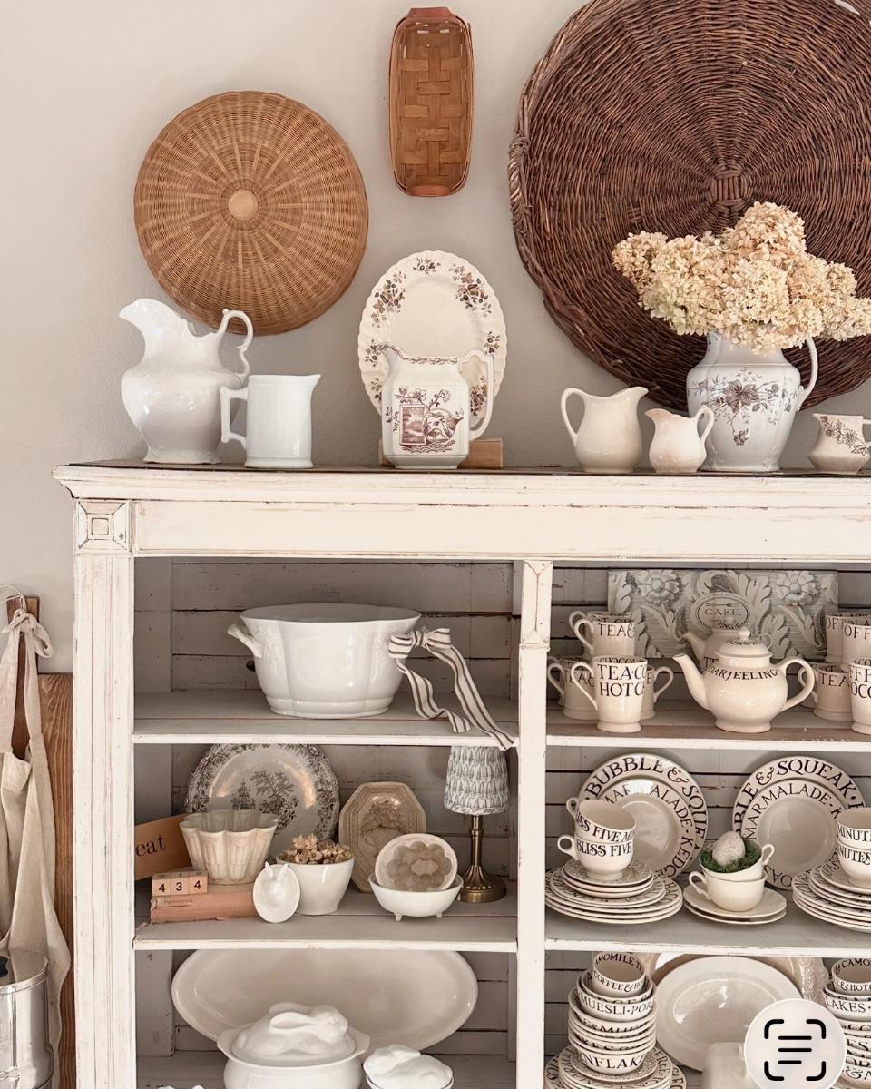 White vintage hutch with assorted cream and brown dishware, pitchers, and bowls. Wicker baskets and dried hydrangeas decorate the display, evoking a cozy, rustic farmhouse style that brings warmth and charm to any home.