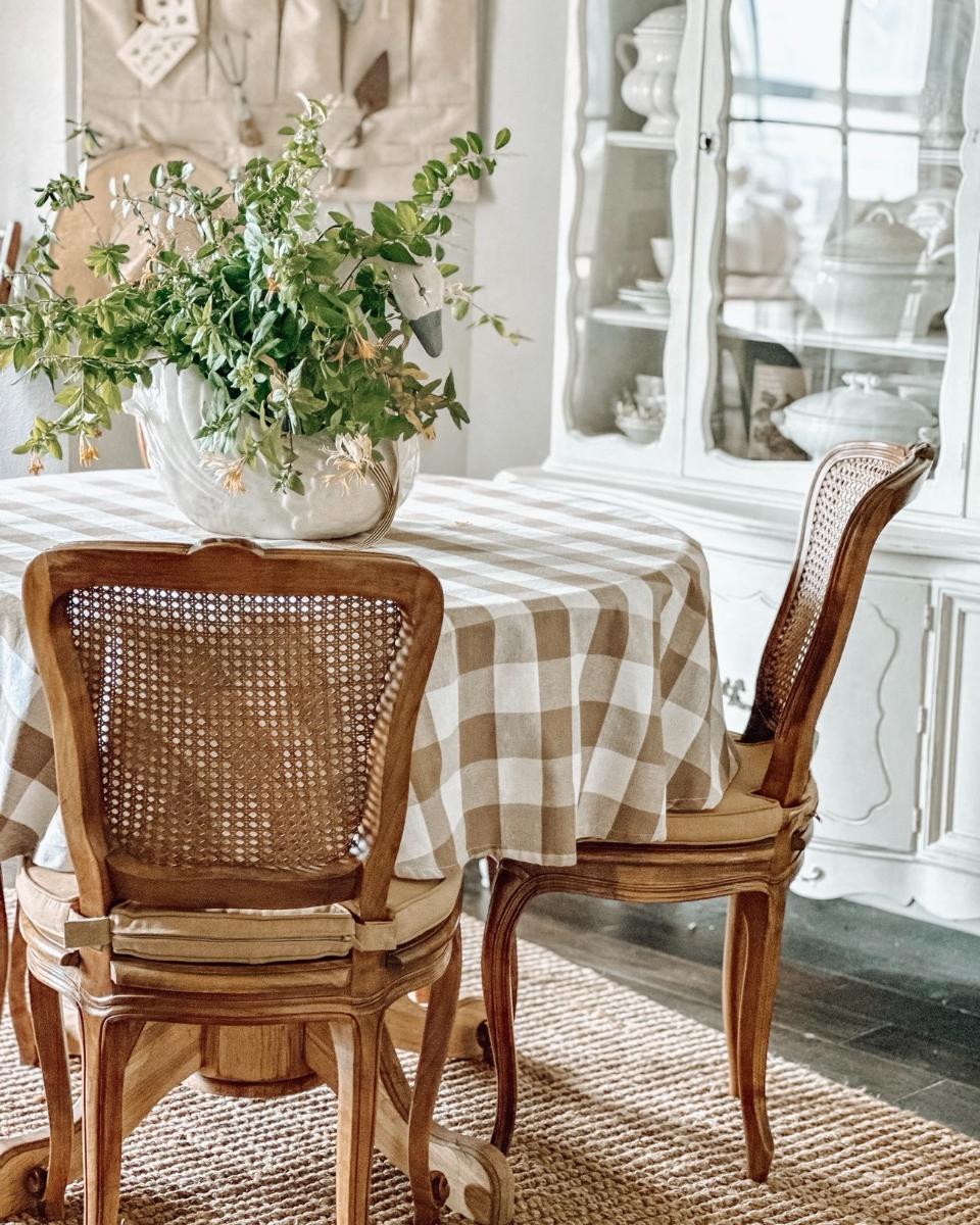 A round table with a beige checkered tablecloth, wooden chairs with woven backs, a large vase of greenery, and a white china cabinet filled with dishes create a cozy, sunlit dining room that feels just like home.