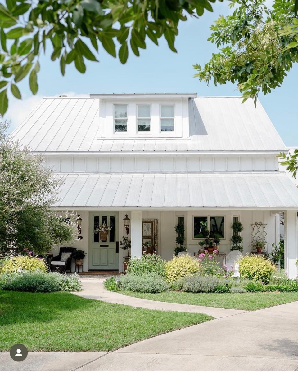 A charming white home with a metal roof, dormer windows, and a cozy front porch is surrounded by lush green bushes, vibrant flowers, and a curved concrete walkway under a bright blue sky.