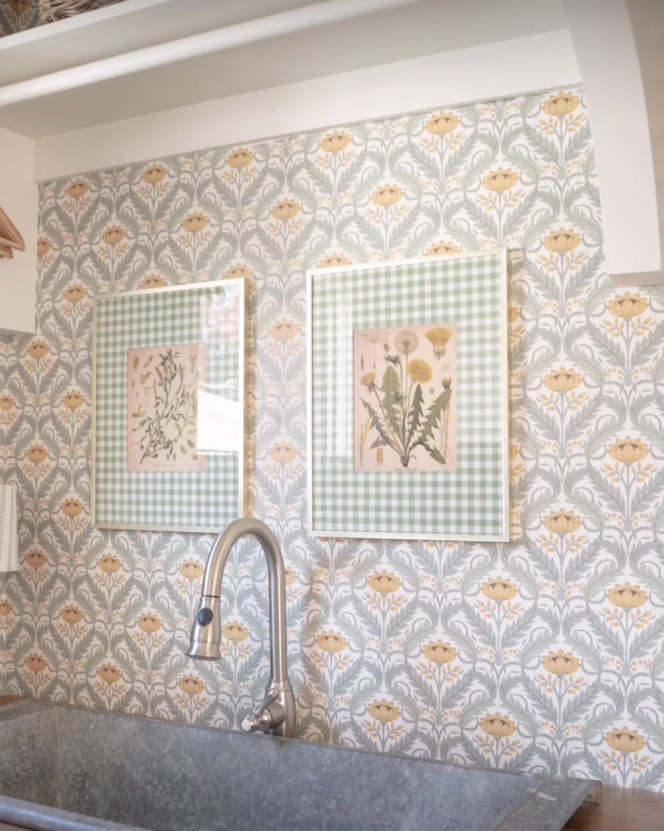 A kitchen sink with a silver faucet is set against a floral-patterned wallpaper, creating a warm and inviting home atmosphere. Above are two framed botanical prints with green checkered borders, featuring illustrations of flowers and plants.