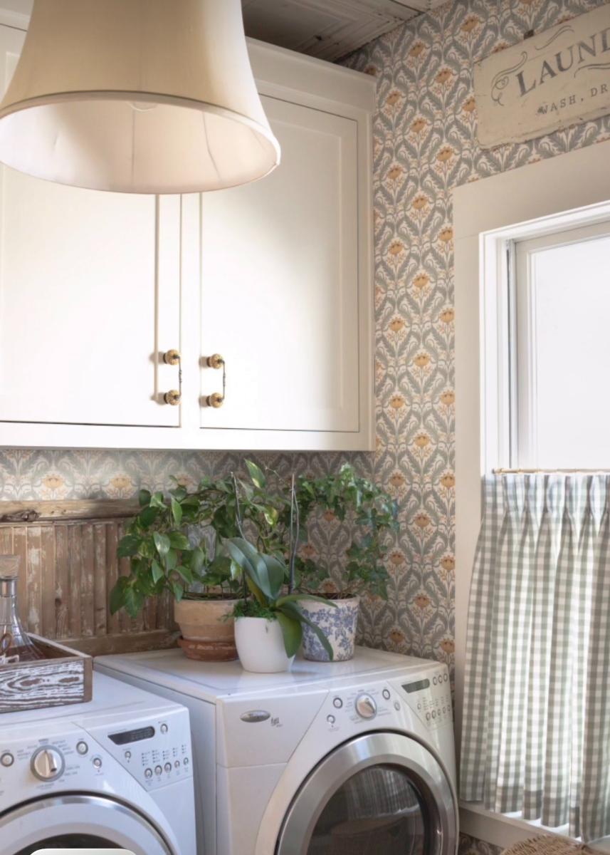 A cozy laundry room brings warmth to your home, featuring patterned wallpaper, white cabinets, potted plants on top of the washer and dryer, a window with checked curtains, and a charming vintage laundry sign.